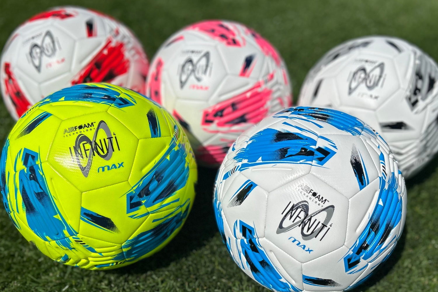Five Samba Infiniti 4 Max Training Balls by Martin Berrill Sports—two white/blue, two white/pink, and one yellow/blue—rest on green grass. Each has precision-moulded grooves for better control.