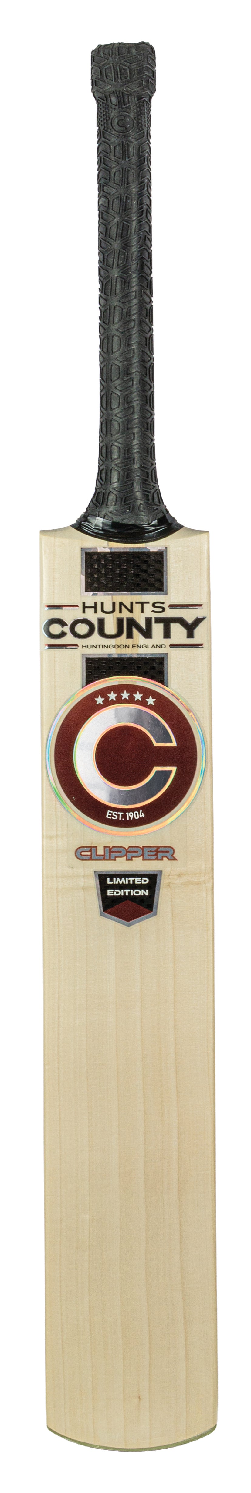 A wooden cricket bat with a black textured grip and Hunts County branding on the blade, featuring a large red and white C logo with additional text and labels below. Crafted from English Willow, the Hunts County Clipper Limited Edition Junior Cricket Bat (2026) is ideal for those seeking quality in junior cricket bats.