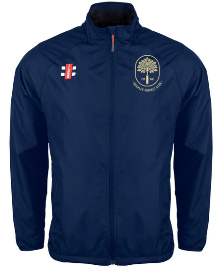 The Gray Nicolls Bushley CC Velocity Rain Jacket is a navy, water-repellent training jacket with long sleeves, high collar, side pockets, a cricket emblem on the left chest, and a red, white, blue logo on the right.