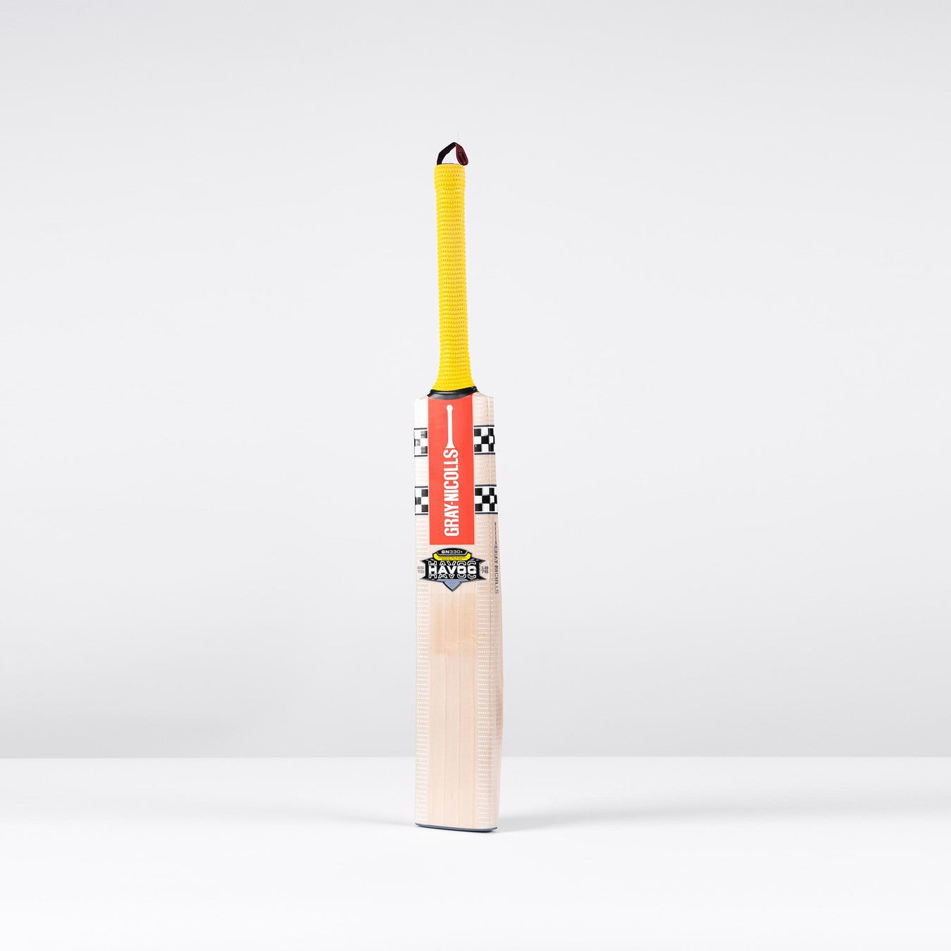 The Gray Nicolls Havoc HB317 GN330+ Junior Cricket Bat 2026 stands upright on a white surface with a yellow grip, orange branding, and checkered black-and-white label, showcasing its ultra-light pickup design against a light grey background.