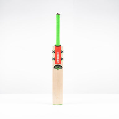 A Gray Nicolls Havoc 1.3 GN130+ Junior Cricket Bat 2026 stands upright on a white surface, featuring a bright green handle and a red front label with the brand name.