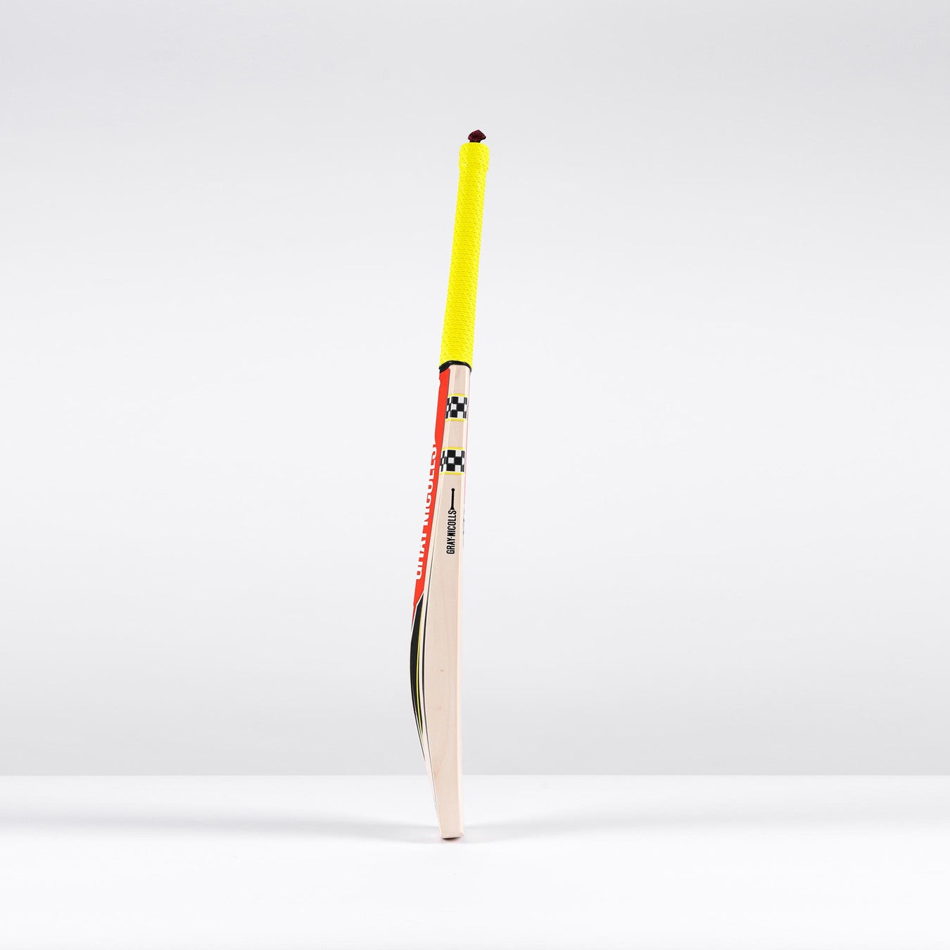 A Gray Nicolls Imperia 1.1 GN330+ Junior Cricket Bat 2026 with a yellow grip stands upright on a white surface against a plain, light gray background.