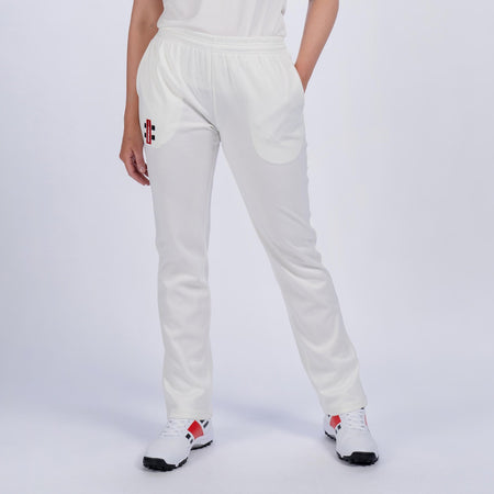 A person models Gray Nicolls Quedgeley & Hardwicke CC Ladies Matrix V2 Cricket Trousers—stylish white joggers with a red and black logo on the left thigh—paired with a white tee and sneakers, against a plain light background.