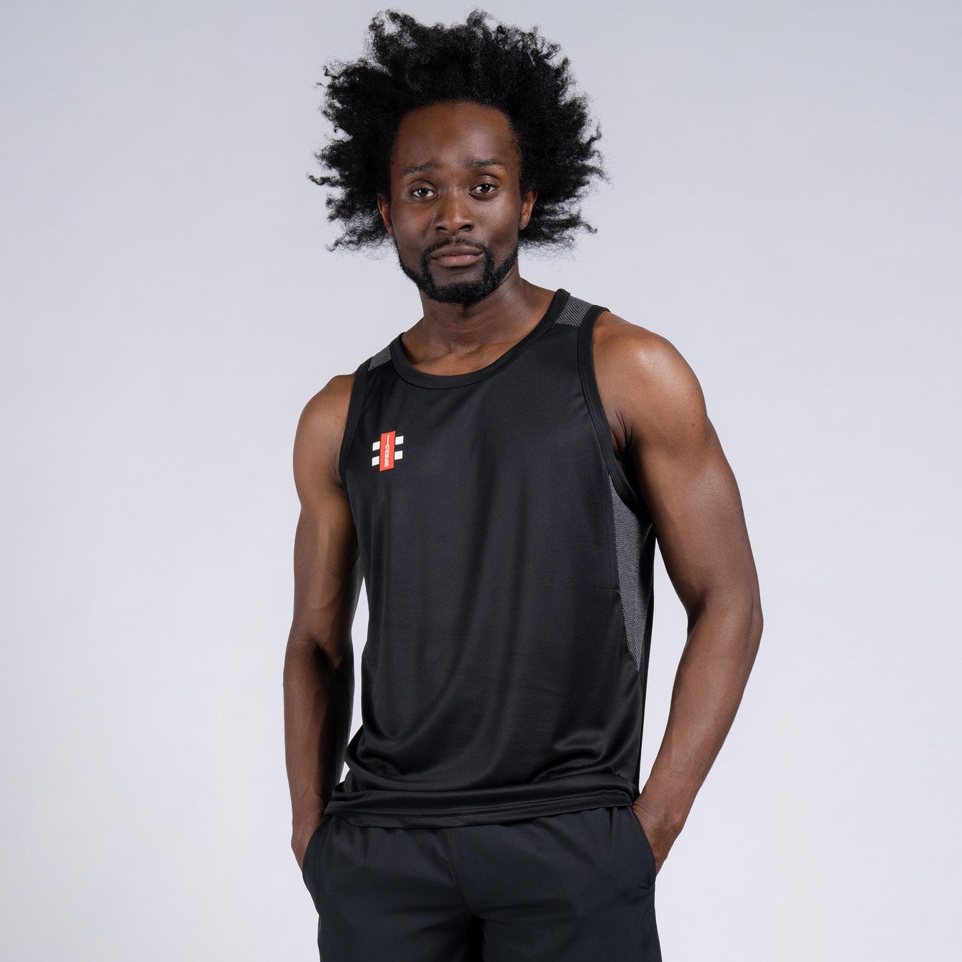 A man with an afro wears the black Gray Nicolls Pro Performance Vest and matching shorts, featuring a small red and white logo on the chest, standing against a plain light grey background.