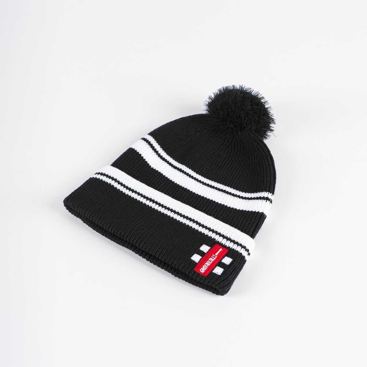 The Gray Nicolls Bobble Hat by Gray Nicolls is black with two white stripes, a black pom-pom on top, and features a red rectangular patch with white text on the folded brim.