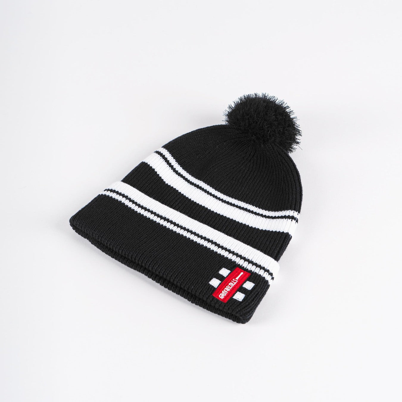The Gray Nicolls Bobble Hat by Gray Nicolls is black with two white stripes, a black pom-pom on top, and features a red rectangular patch with white text on the folded brim.