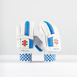 A pair of Gray Nicolls Havoc 1.0 700 Batting Gloves 2026, white and blue with checkered wristbands and HAVOC branding, featuring enhanced finger protection, stand upright on a white surface against a plain light gray background.