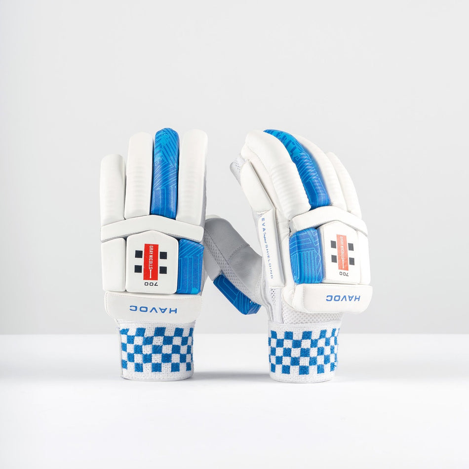 A pair of Gray Nicolls Havoc 1.0 700 Batting Gloves 2026, white and blue with checkered wristbands and HAVOC branding, featuring enhanced finger protection, stand upright on a white surface against a plain light gray background.