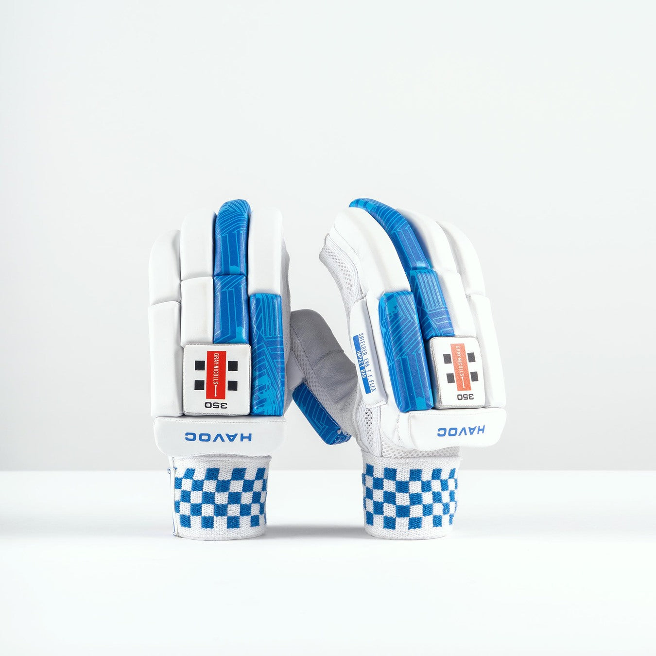 A pair of Gray Nicolls Havoc 1.0 350 Batting Gloves 2026 in white with blue accents and checkered wristbands stand upright on a white surface, showing finger protection and the Gray Nicolls brand name on the wrist.