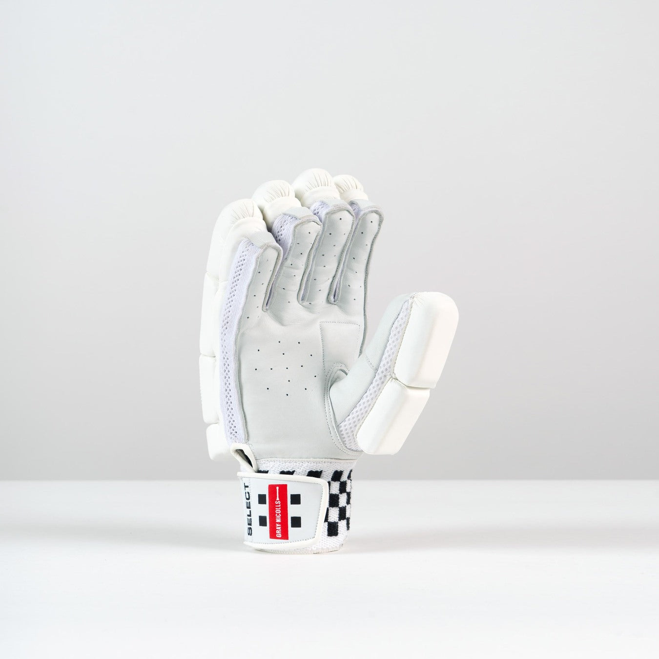 The Gray Nicolls Classic Select Batting Gloves 2026 stand upright on a light gray background, showcasing white padding, finger protection, perforations, mesh accents, a leather palm, and a wrist strap with a red and black checkered label.