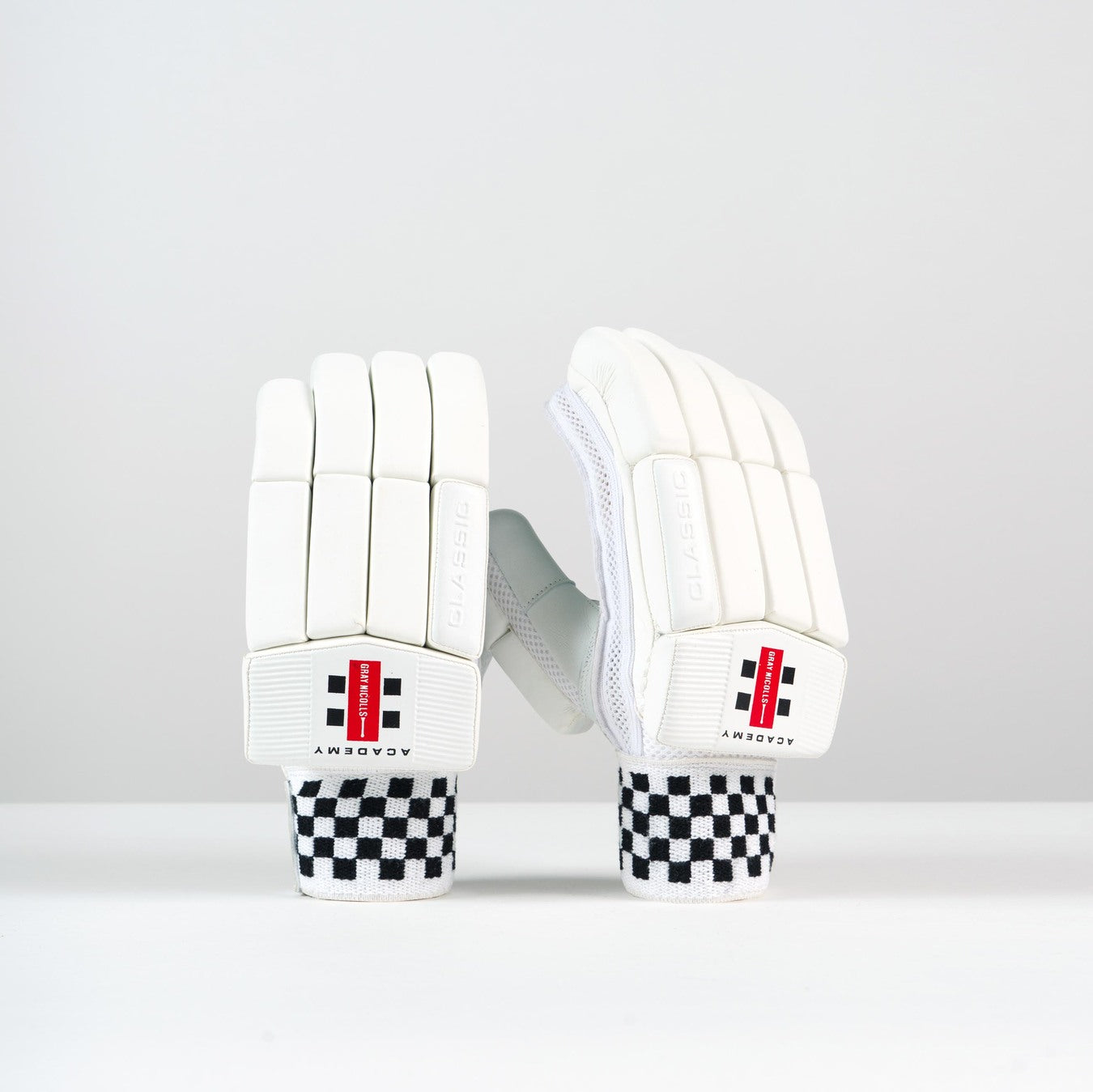 The Gray Nicolls Classic Academy Batting Gloves 2026, with a leather palm, black and white checkered cuffs, and red, black, and white branding on the wrists, are displayed upright against a plain white background.