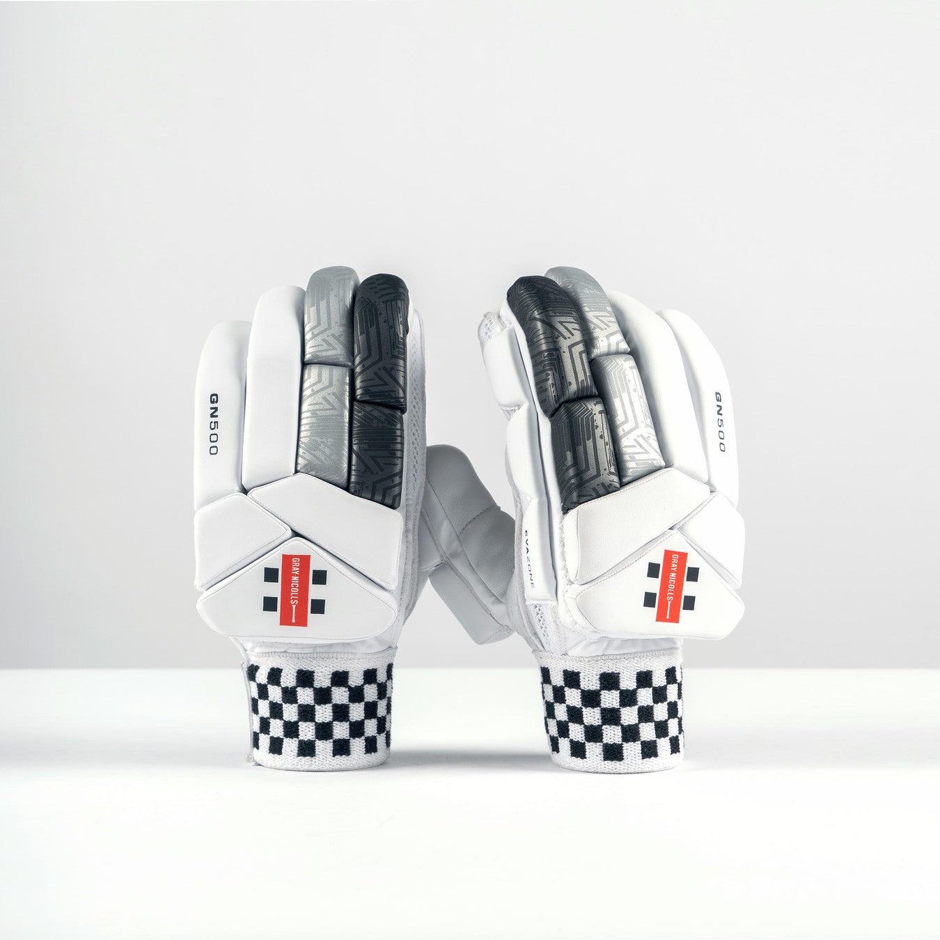 A pair of Gray Nicolls GN500X Batting Gloves 2026 featuring white, black, and gray high-density foam padding with black-and-white checkered wrist straps and red rectangular accents, displayed upright on a plain white surface.