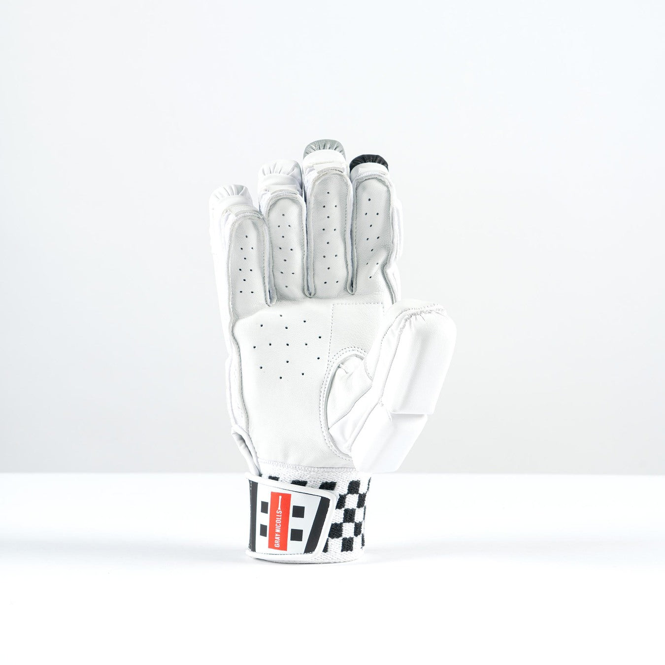 The Gray Nicolls GN500X Batting Glove 2026 is a white, lightweight glove with perforations and high-density foam padding. It stands upright on a white background, featuring a red and black wrist label with a black-and-white checkered pattern.