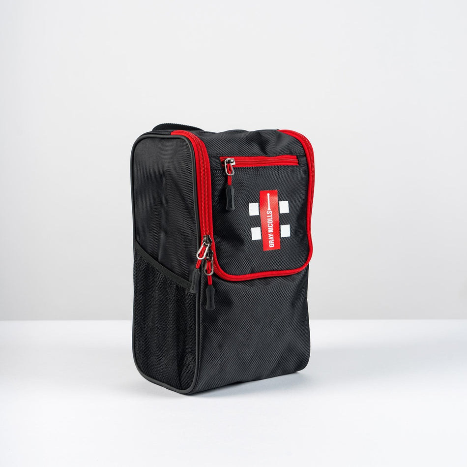 The Gray Nicolls Team Shoe Bag 2026 by Gray Nicolls is a black, rectangular cricket shoe bag with red trim, white cross symbols, and a red EMERGENCY label. It has a zippered front pocket, mesh side pocket, and is shown on a white surface.