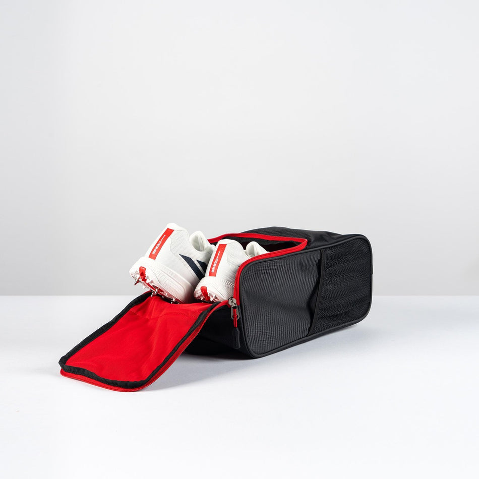 A black and red Gray Nicolls Team Shoe Bag 2026 by Gray Nicolls sits on a white surface with its zipper open, showing a pair of white and red sneakers stored inside.