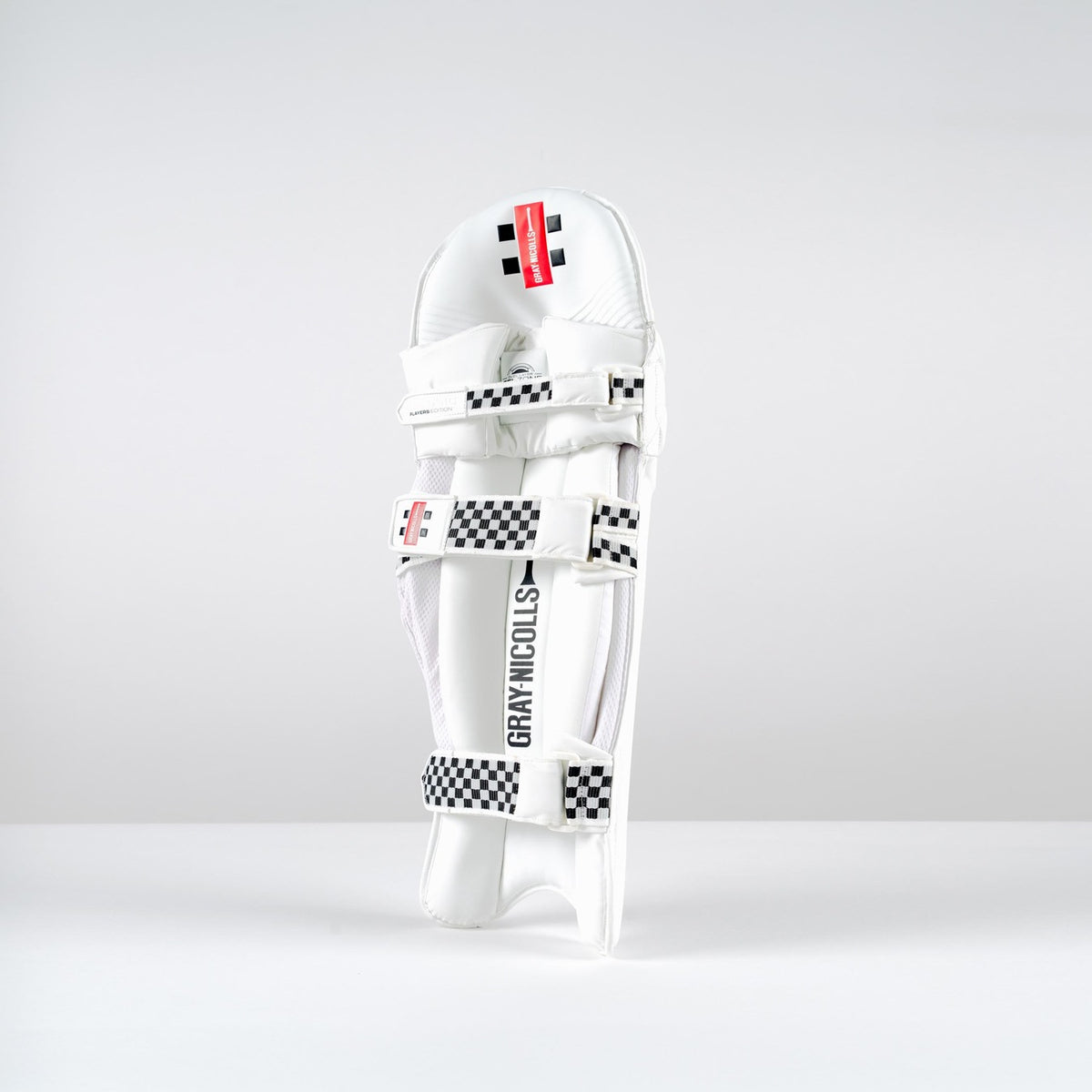 A Gray Nicolls Classic Players Edition Batting Pad 2026 stands upright on a white surface, featuring black-and-white checkered straps, a red label, and lightweight HDF multilayer protection against a plain light gray background.