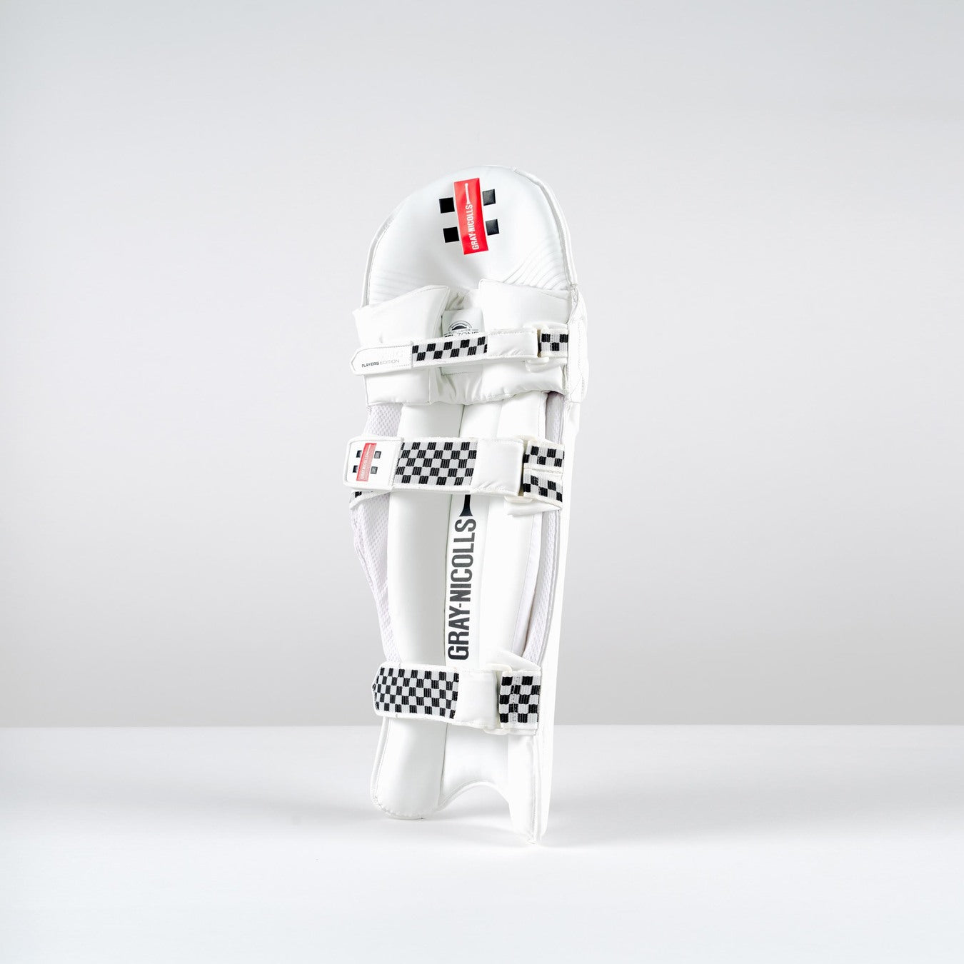 A Gray Nicolls Classic Players Edition Batting Pad 2026 stands upright on a white surface, featuring black-and-white checkered straps, a red label, and lightweight HDF multilayer protection against a plain light gray background.