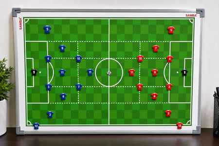The Samba Sports Football Coaching Board Shirt Magnets Set of 27 displays a soccer field layout with blue and red team formations, each represented by numbered magnetic shirt markers, along with two black goalkeeper magnets and a central ball. The coaching board is shown on a desk beside a plant and pen holder.