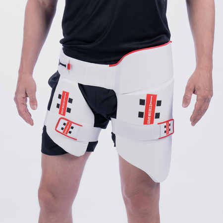 A person wearing a Gray Nicolls All in One 360 Thigh Pad, featuring white padding with red accents and brand labels, over black shorts and a black shirt.