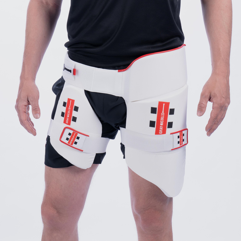 A person wearing a Gray Nicolls All in One 360 Thigh Pad, featuring white padding with red accents and brand labels, over black shorts and a black shirt.