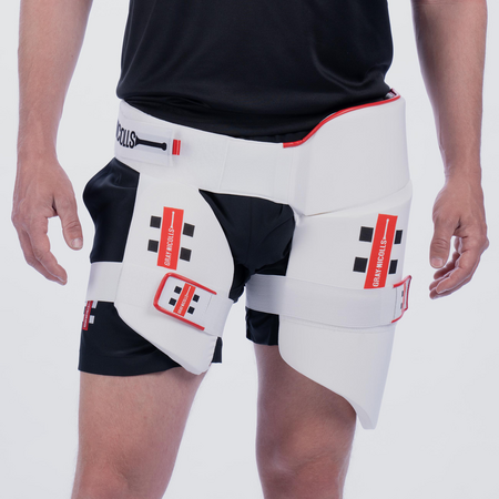 A person wears black shorts and a Gray Nicolls All in One 360 Thigh Pad—a white, adjustable thigh guard with red accents for cricket, designed for injury prevention and support.