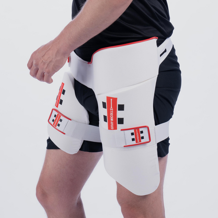 A person is shown from the side against a plain light background wearing the Gray Nicolls All in One 360 Thigh Pad with red and black details over black shorts.