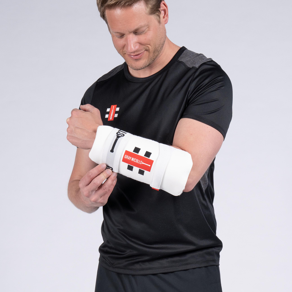 A man in a black sports shirt adjusts the Gray Nicolls Test Armguard with red and black accents on his left arm, smiling slightly as he looks down at it against a plain light gray background.