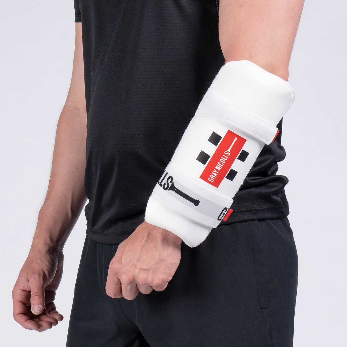 A person in black attire is shown from the shoulders down, wearing a Gray Nicolls Test Armguard—featuring dual-level protection with white padding, black squares, and a red stripe—secured around their right forearm and elbow.