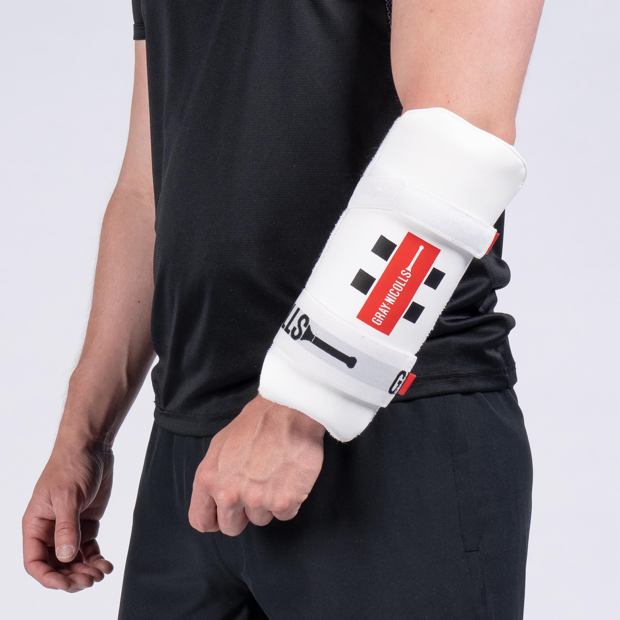 A person in black attire is shown from the shoulders down, wearing a Gray Nicolls Test Armguard—featuring dual-level protection with white padding, black squares, and a red stripe—secured around their right forearm and elbow.