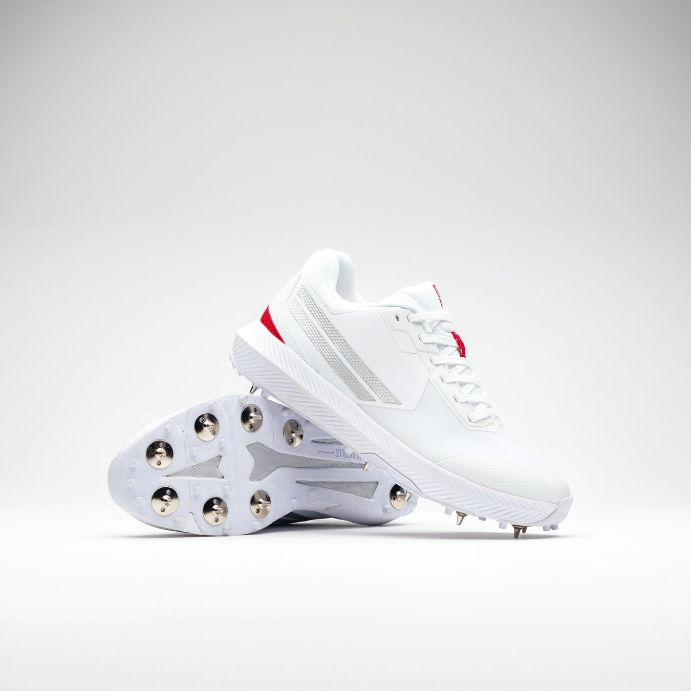 The Gray Nicolls Revo Pro 2.0 Cricket Spikes by Gray Nicolls feature white uppers with silver and red accents, a Maxcool Duo upper, Energise midsole, and metal studs. One shoe stands upright while the other shows the studded sole.