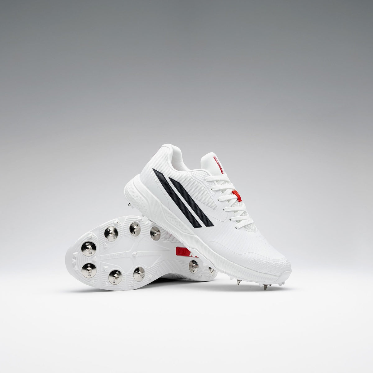 A pair of Gray Nicolls Revo Pro 3 Cricket Spikes is displayed against a gradient background, one upright and one leaning to highlight the metal spikes, featuring lightweight design with black stripes and red accents.