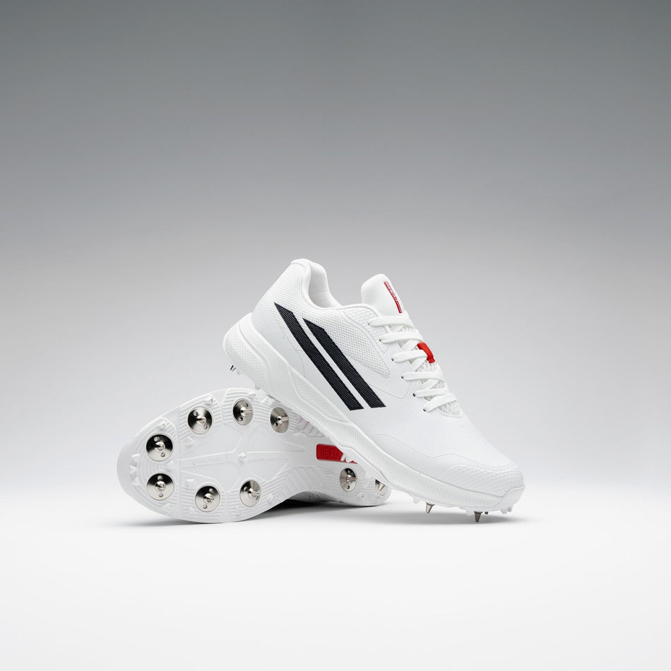 A pair of Gray Nicolls Revo Pro 3 Cricket Spikes is displayed against a gradient background, one upright and one leaning to highlight the metal spikes, featuring lightweight design with black stripes and red accents.