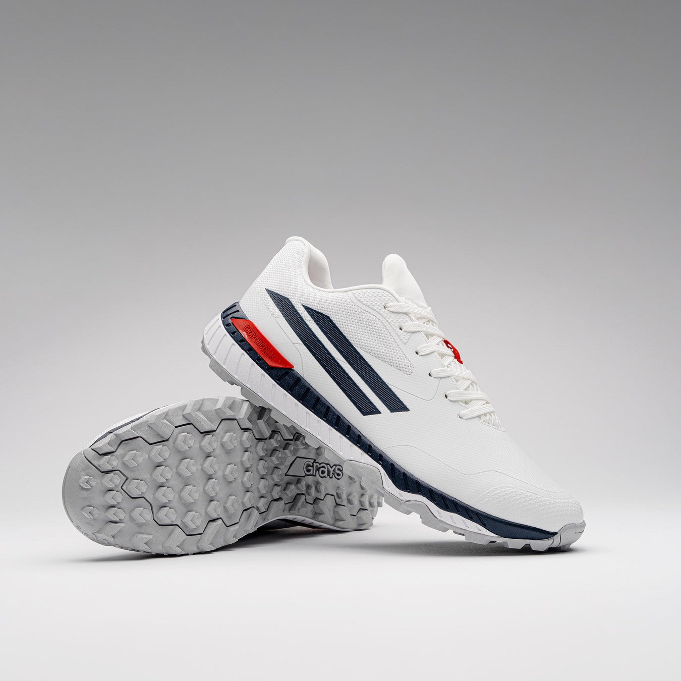 The Gray Nicolls Revo Pro 3 Cricket Batting Shoe features a white upper with dark blue stripes, red accents, and textured gray soles. One shoe stands upright while the other lies on its side to display the tread pattern.