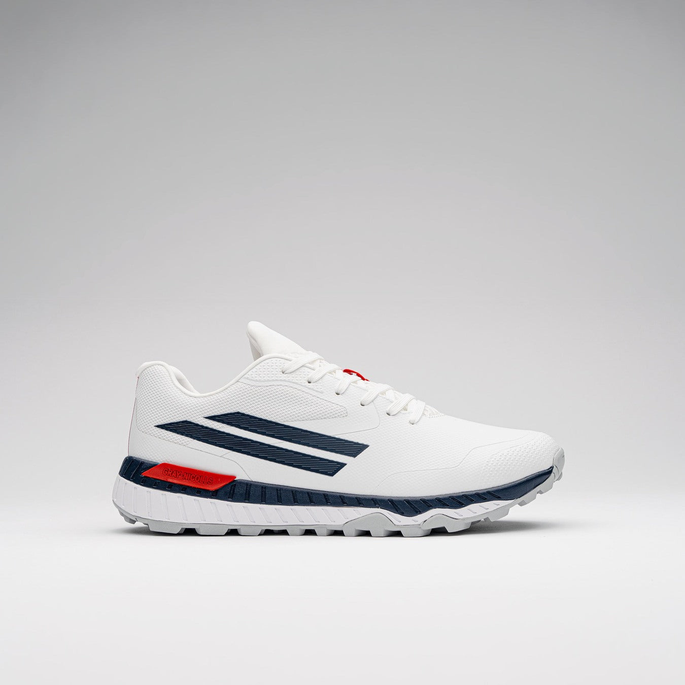 The Gray Nicolls Revo Pro 3 Cricket Batting Shoe by Gray Nicolls is a white athletic shoe with dark blue stripes, red heel accent, and textured white sole, featuring a lightweight design and shown on a plain light gray background.