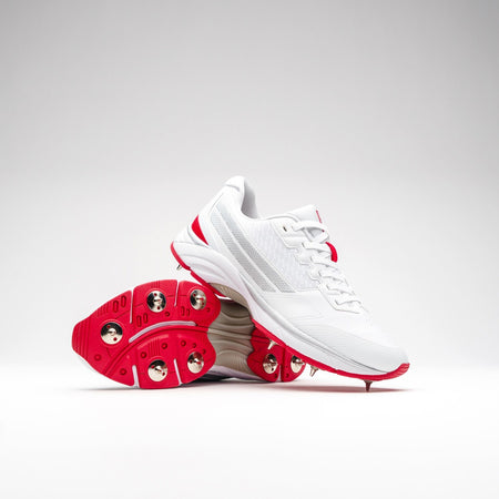 The Gray Nicolls Velocity 5.0 Cricket Spikes, featuring white uppers with red accents and metal sole spikes, are shown on a light background—one upright and one tilted to display the spike design.