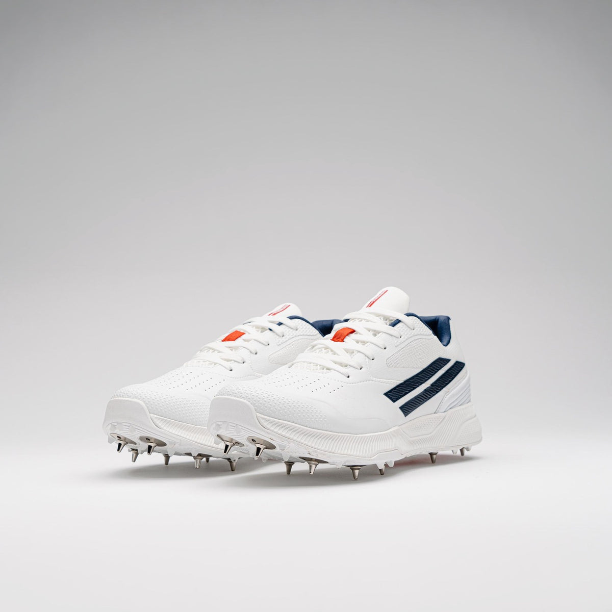 The Gray Nicolls Players 4.0 Cricket Spikes are premium white cricket shoes with metal spikes, navy blue stripes, and orange accents by Gray Nicolls, shown on a light gray background.