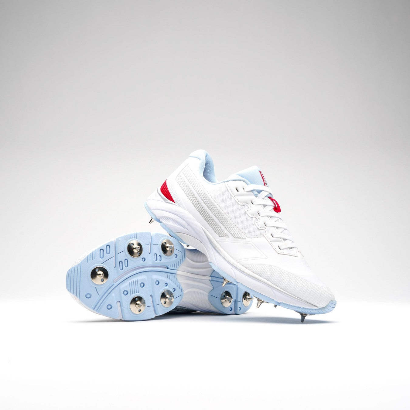 Gray Nicolls Velocity 5.5 Narrow Cricket Spikes for women, shown on a white background with one upright and one displaying metal spikes. Features white uppers, light blue soles, and red accents—perfect for women cricketers needing a narrow fit.