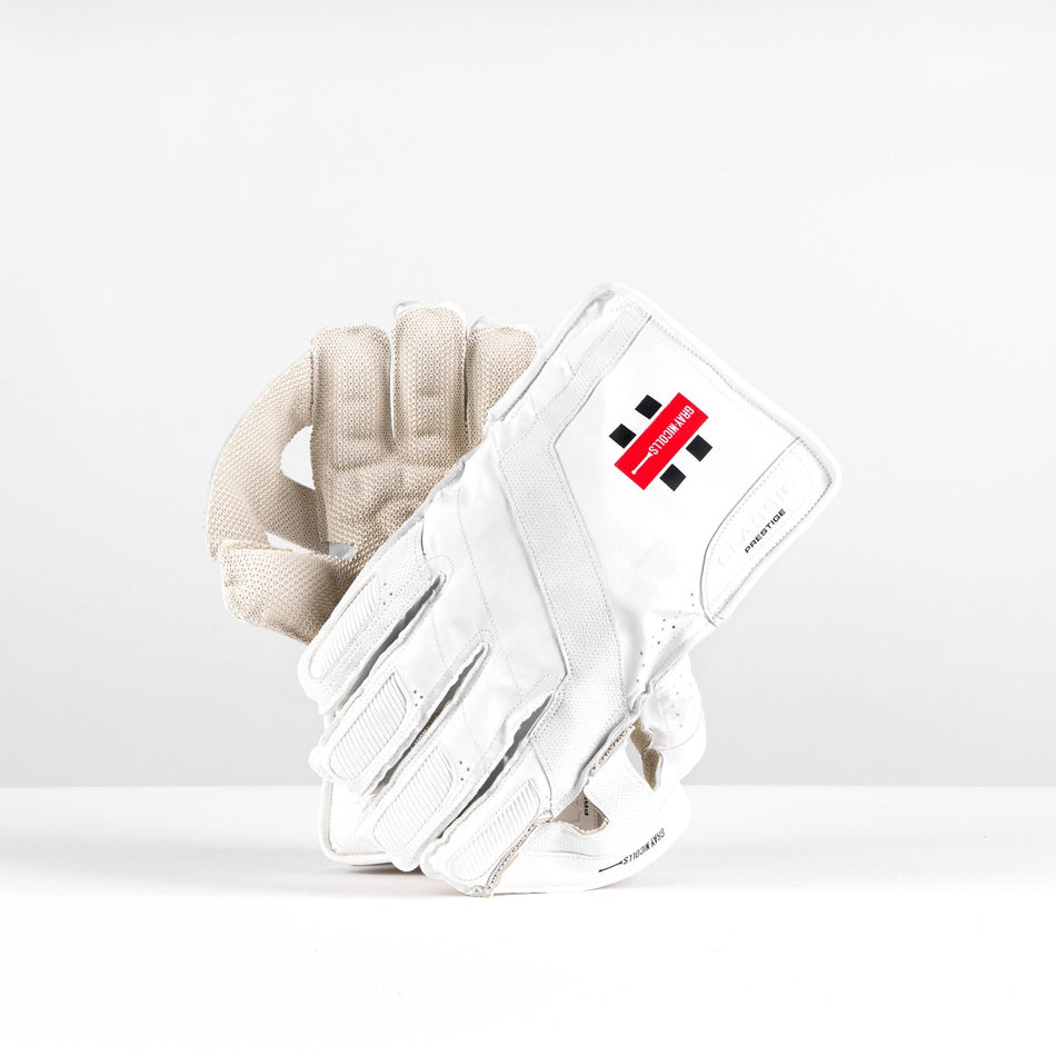 The Gray Nicolls Classic Prestige WK Gloves 2026 feature padded fingers for impact protection and a red and black logo on the wrist. This white pair is displayed upright on a white surface with a plain white background.