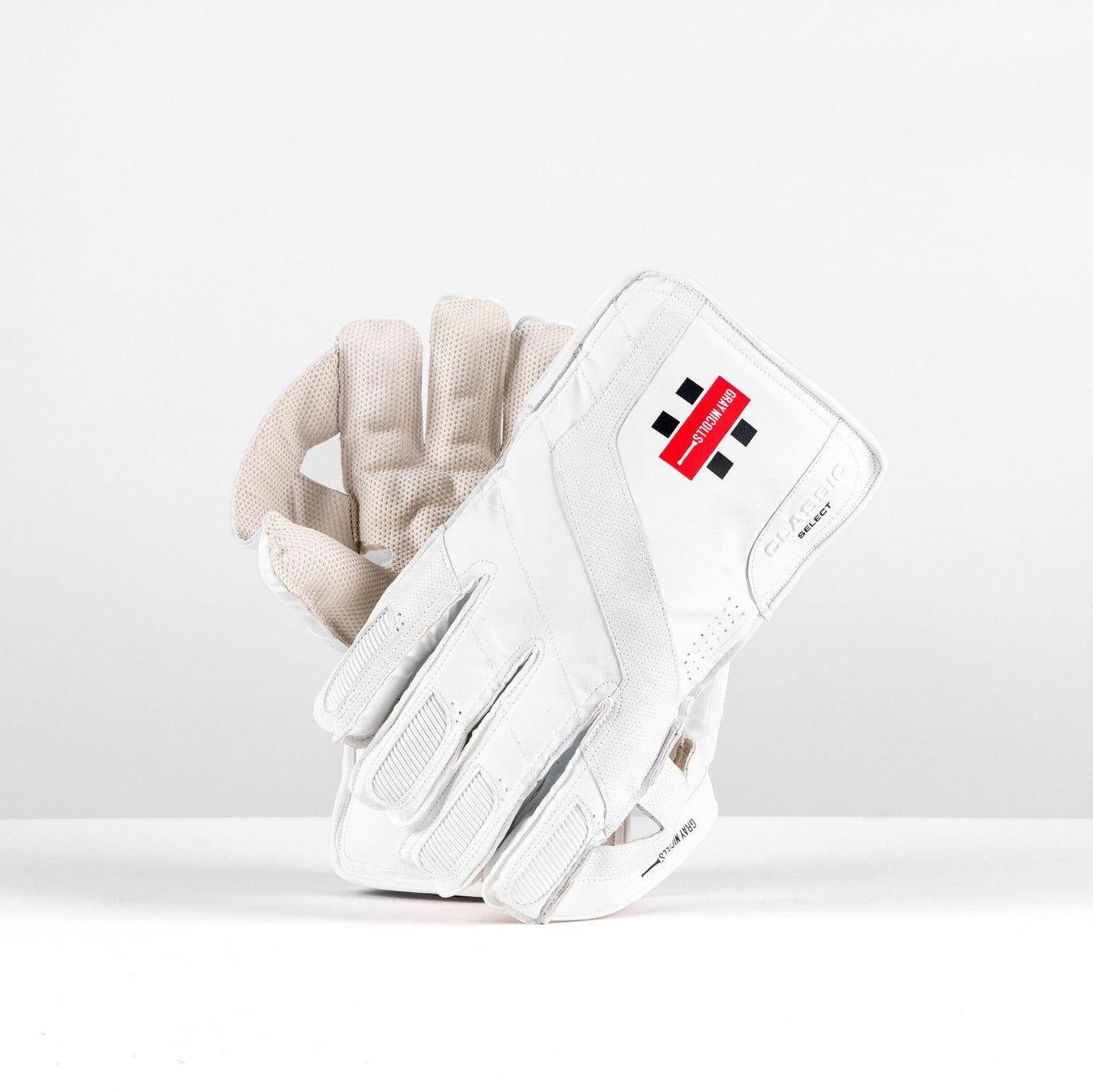 The Gray Nicolls Classic Select WK Gloves 2026 for juniors are white wicket keeping gloves with padded fingers, a red and black logo on the wristband, and a ping pong grip, displayed upright on a plain white background.
