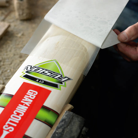 A person applies a Gray Nicolls Extratec self-adhesive PU facing to a cricket bat blade for added protection, securing it on a workbench.