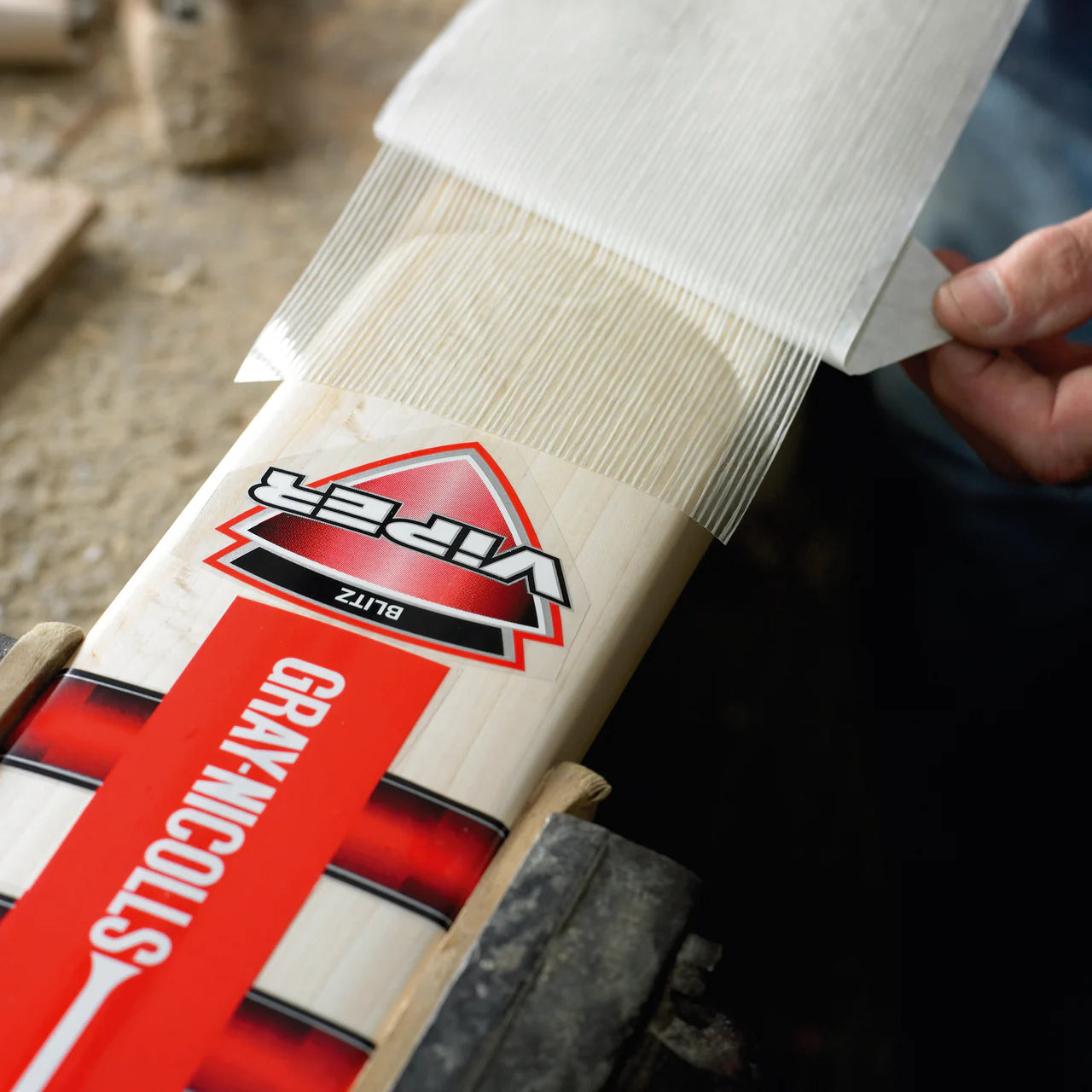 A person applies a Gray Nicolls Fibretec Sheet to a Viper cricket bat for extra protection, securing it on a workbench.