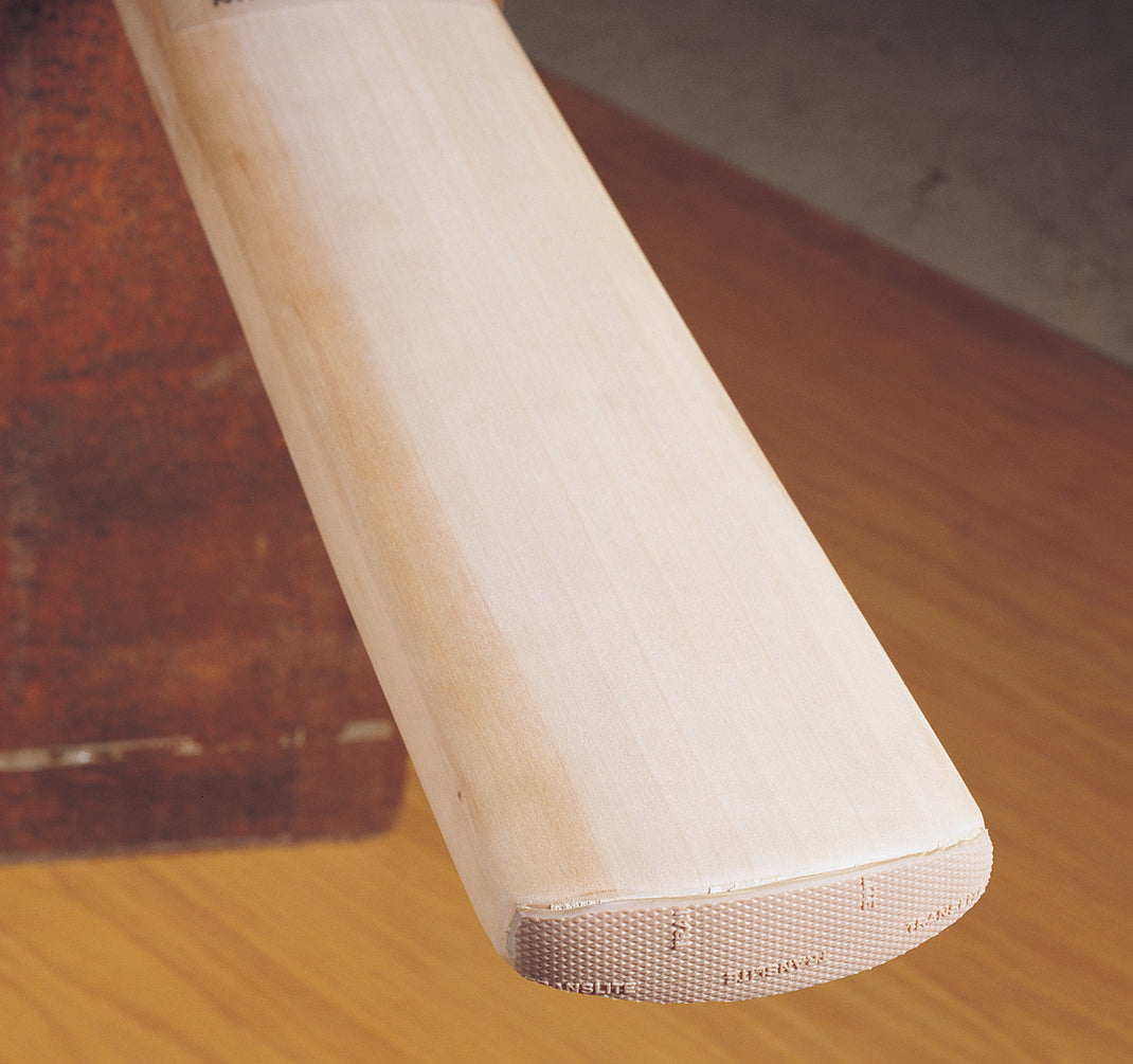 Close-up of a wooden cricket bat with a smooth, light-colored face and textured end, featuring a Gray-Nicolls Protectoe toe guard by Gray Nicolls for enhanced protection, resting on a brown surface.