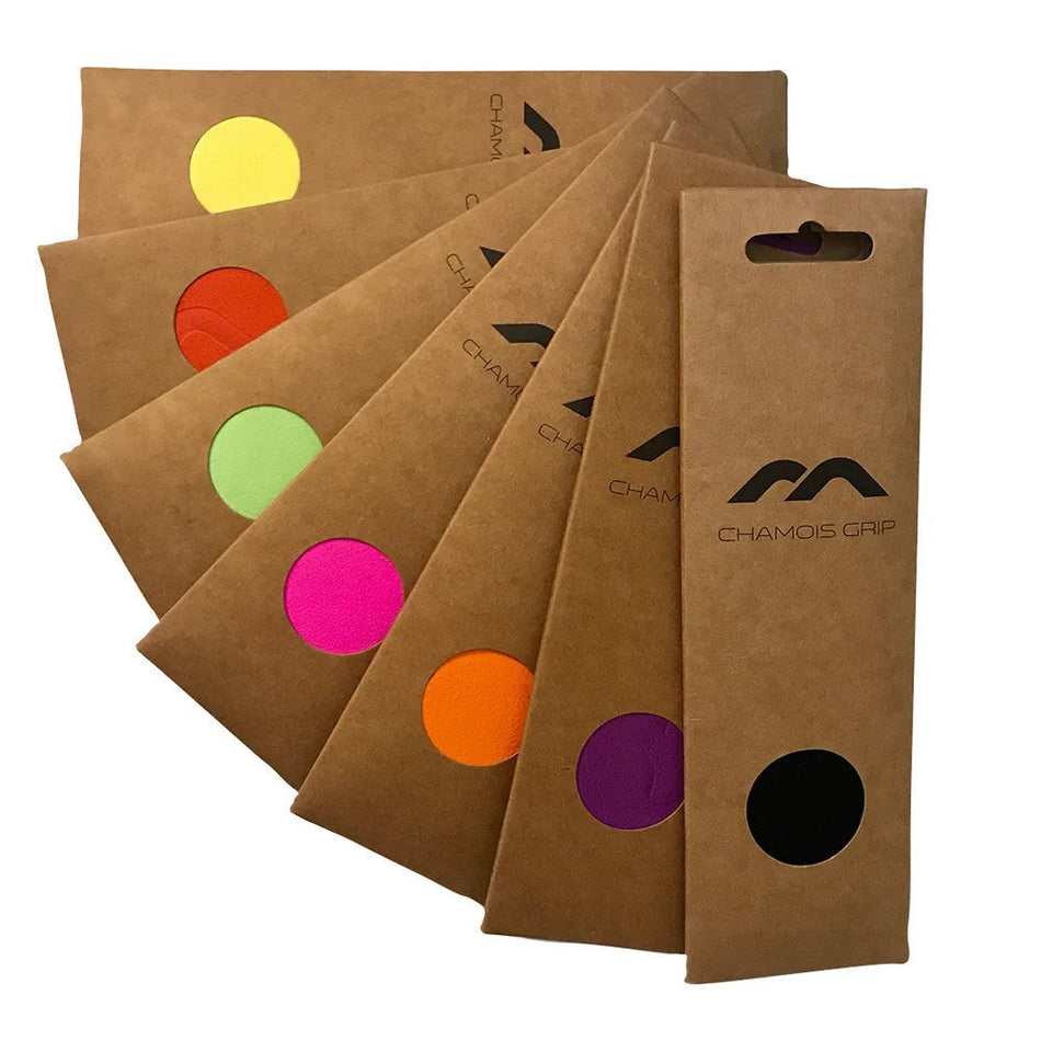 Ten brown Mercian Chamois Hockey Grip packs are fanned out, each showing a circular grip in yellow, red, green, pink, orange, or black. Each pack displays the Mercian logo and contains one hockey stick grip.
