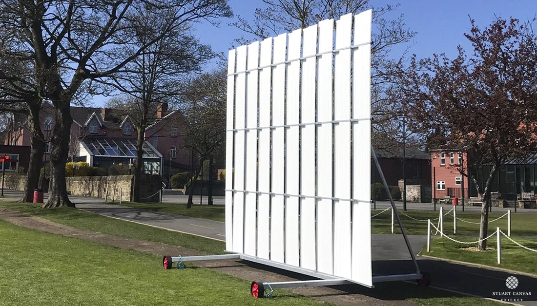The Stuart Canvas Polypropylene Sightscreen, finished in white and mounted on wheels for easy movement, stands on a grassy field near a path with houses, trees, and blue sky in the background.