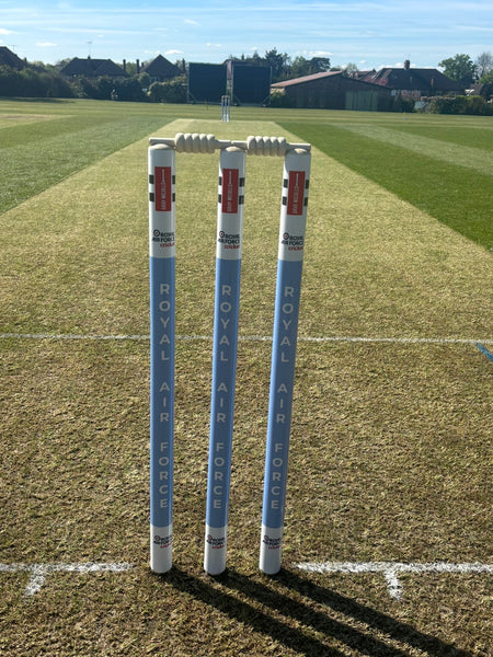 A close-up of Gray Nicolls Custom Printed Club Cricket Stumps on a well-kept pitch under sunny skies, featuring "Royal Air Force" text, with a scoreboard and trees visible in the background.