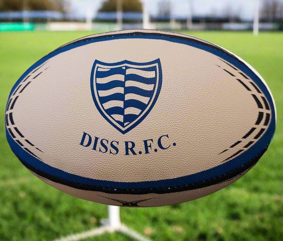 A close-up of a Custom Gilbert Match or Training Rugby Ball with logo, featuring a blue and white shield and DISS R.F.C. text, lying on grass with rugby goalposts and trees in the background.