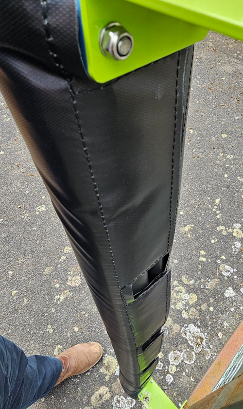 Close-up of Ritelite Sports Lite Fence Kit Padding in black, stitched and bolted to a green metal post for player protection. A person in jeans and tan boots stands nearby on asphalt.