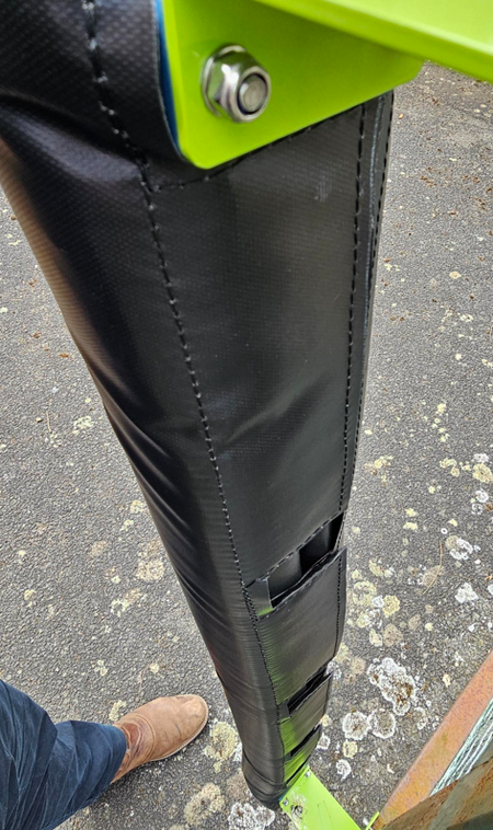 Close-up of Ritelite Sports Lite Fence Kit Padding in black, stitched and bolted to a green metal post for player protection. A person in jeans and tan boots stands nearby on asphalt.