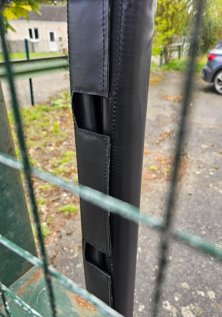 A close-up of Ritelite Sports Lite Fence Kit Padding in black, securely attached to a metal post for enhanced player protection, with a blurred background featuring a parked car, building, and greenery.
