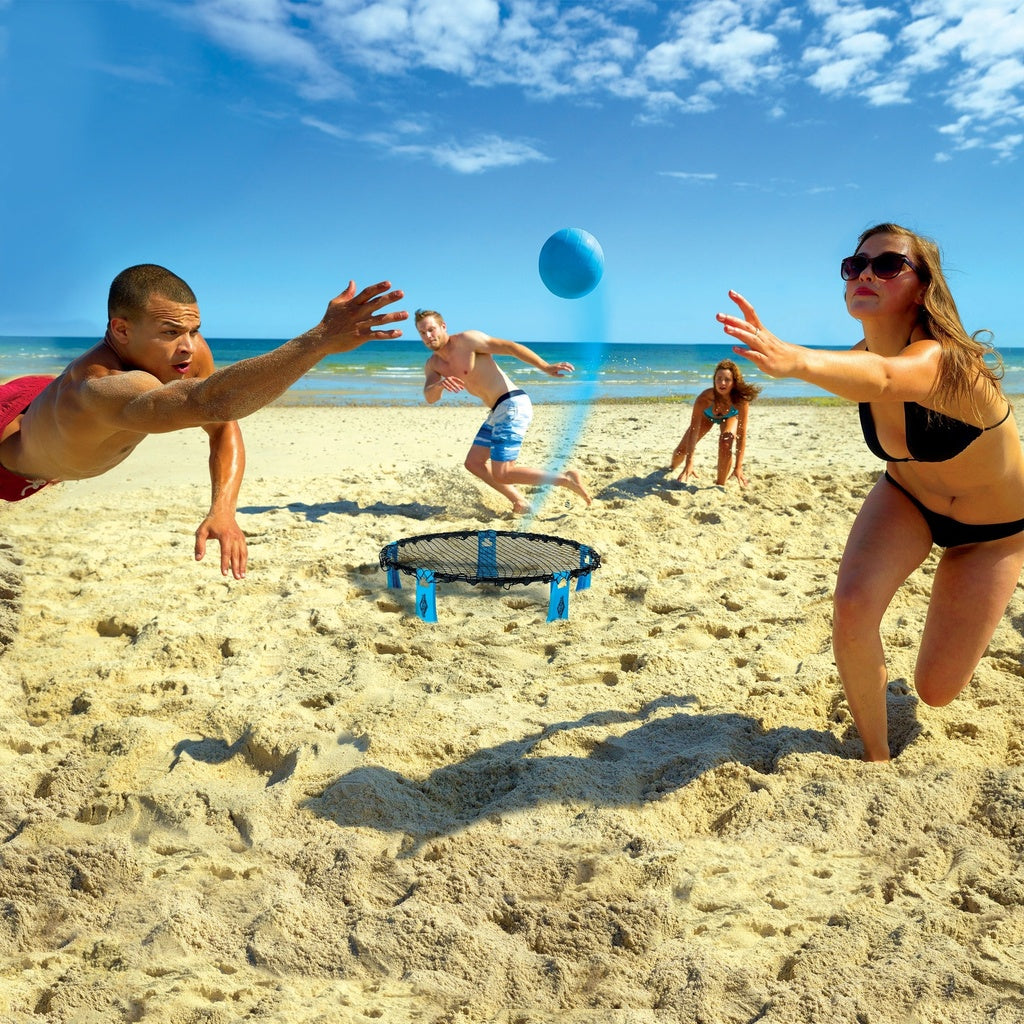 Four people play Franklin Spyderball by Franklin on a sandy beach under a blue sky. Two players dive for the ball while teammates look on, with the ocean in the background. This versatile outdoor game adds excitement to any outing.